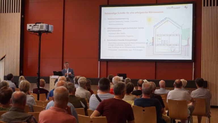 Das Publikum ist von hinten in einem Saal zu sehen. Vorn steht ein Mann leicht links der Mitte und hält ein Mikrofon in der Hand. Rechts oben ist eine große Leinwand mit einer Präsentation zu erkennen.