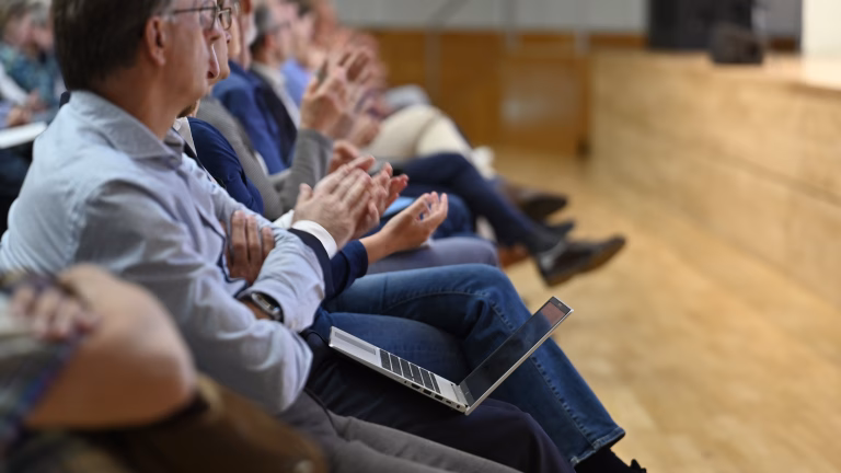 Das Publikum in der ersten Reihe sitzt seitlich, alle klatschen und blicken nach oben rechts. Die Köpfe sind entsprechend zur Seite gedreht, die meisten sitzen mit überkreuzten Beinen.