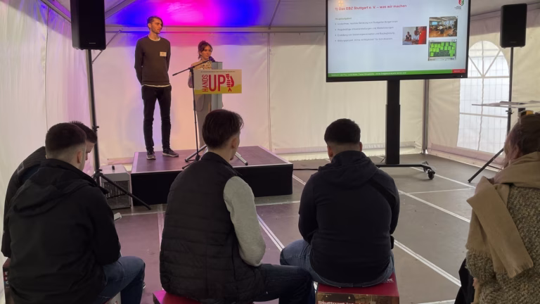 Unten mittig sitzen drei Personen auf bedruckten Klötzen, man sieht sie nur von hinten. Oben mittig links stehen zwei Mitarbeitende des EBZ, ein Mann und eine Frau, auf einer Bühne. Die Frau spricht ins Mikrofon. Rechts befindet sich ein großer Bildschirm, auf dem das EBZ vorgestellt wird.