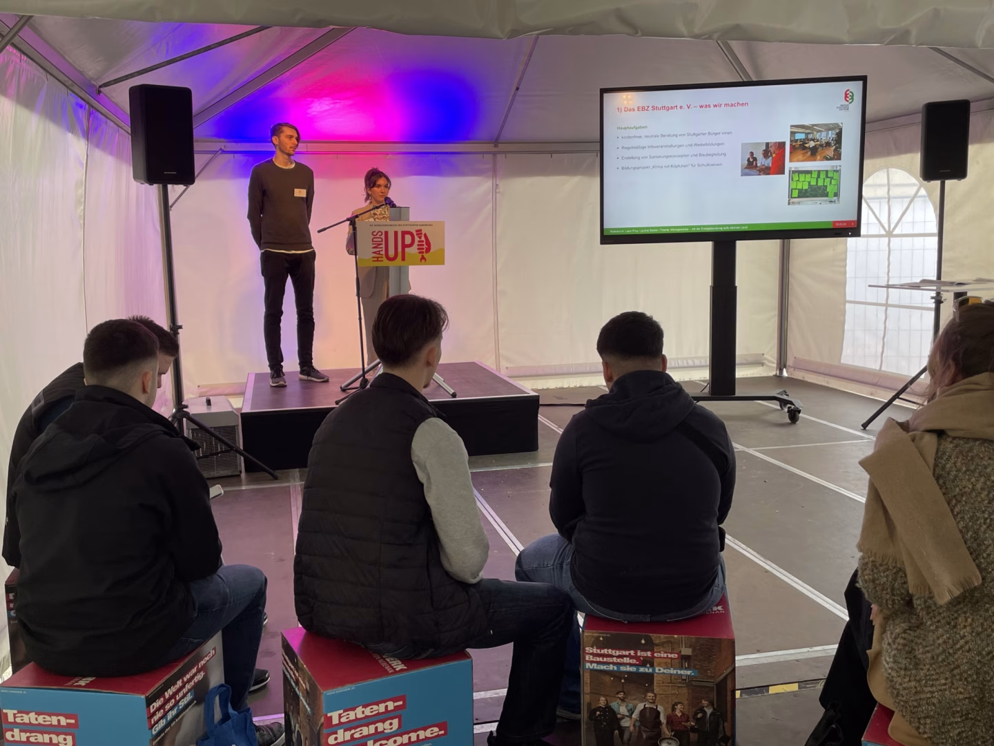 Unten mittig sitzen drei Personen auf bedruckten Klötzen, man sieht sie nur von hinten. Oben mittig links stehen zwei Mitarbeitende des EBZ, ein Mann und eine Frau, auf einer Bühne. Die Frau spricht ins Mikrofon. Rechts befindet sich ein großer Bildschirm, auf dem das EBZ vorgestellt wird.