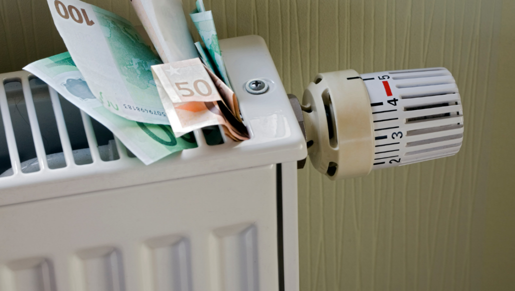 Detailaufnahme einer Heizung mit sichtbarem Regler. Auf dem oberen Bereich der Heizung stecken mehrere 100- und 50-Euro-Scheine.