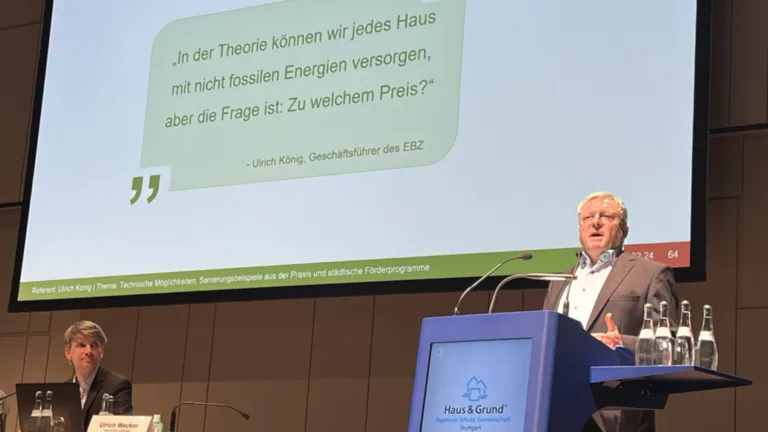 Der ehemalige Geschäftsführer des EBZ steht auf einer Bühne und spricht in ein Mikrofon. Hinter ihm ist eine Leinwand mit einer PowerPoint-Präsentation zu sehen.