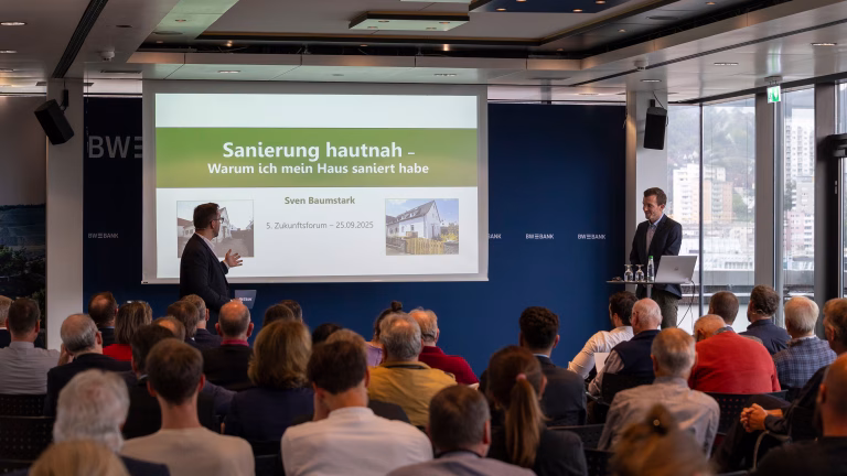 Zwei Männer stehen vor dem Publikum und besprechen eine Präsentationsfolie auf der steht "Sanierung hautnah - warum ich mein Haus saniert habe"