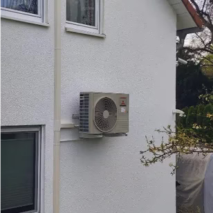 Ein quaderförmiges Außengerät einer Wärmepumpe oder Klimaanlage ist an einer weißen Hausfassade montiert. Rechts daneben sind einige Äste eines Strauchs im Vordergrund zu sehen.