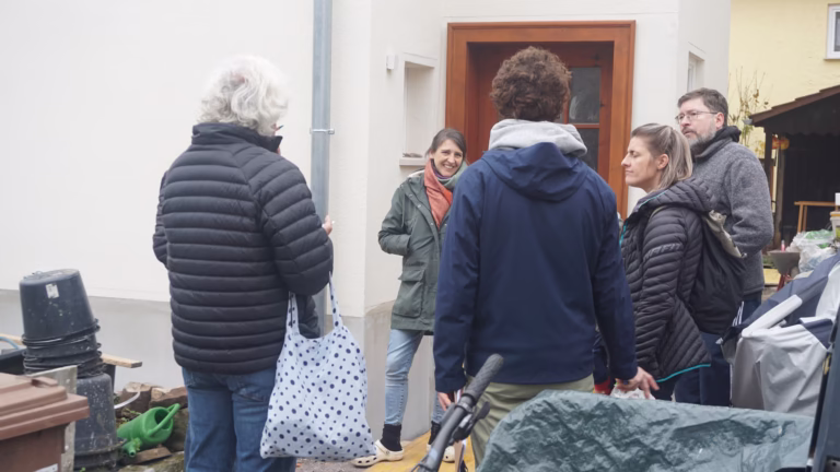 Menschen stehen in einer Gruppe vor einem Haus und unterhalten sich, eine Frau lacht freundlich